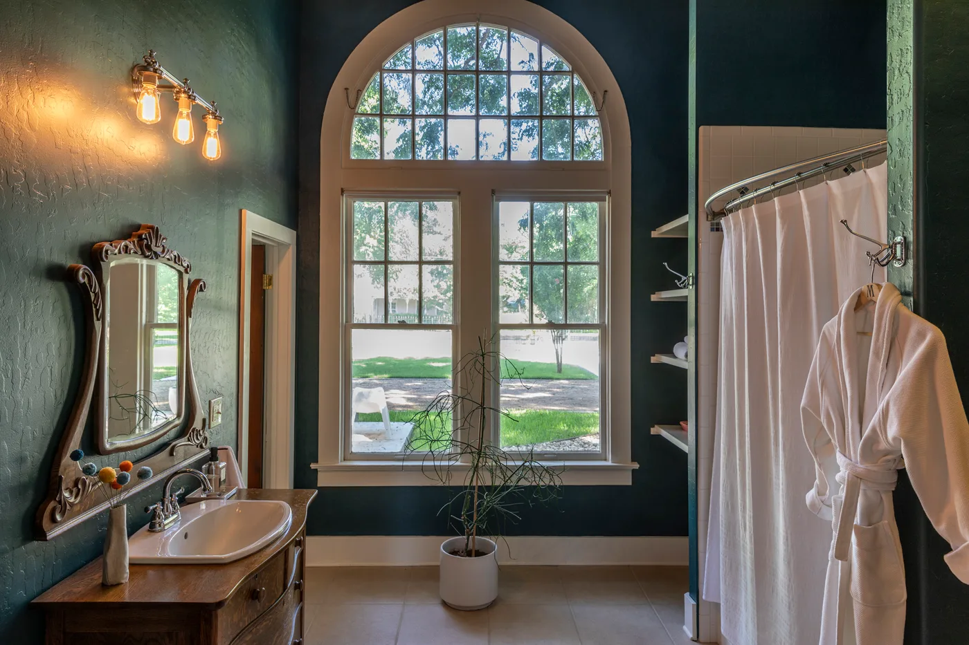 Deep teal bathroom with tall arched window, carved antique mirror and white robe by the shower