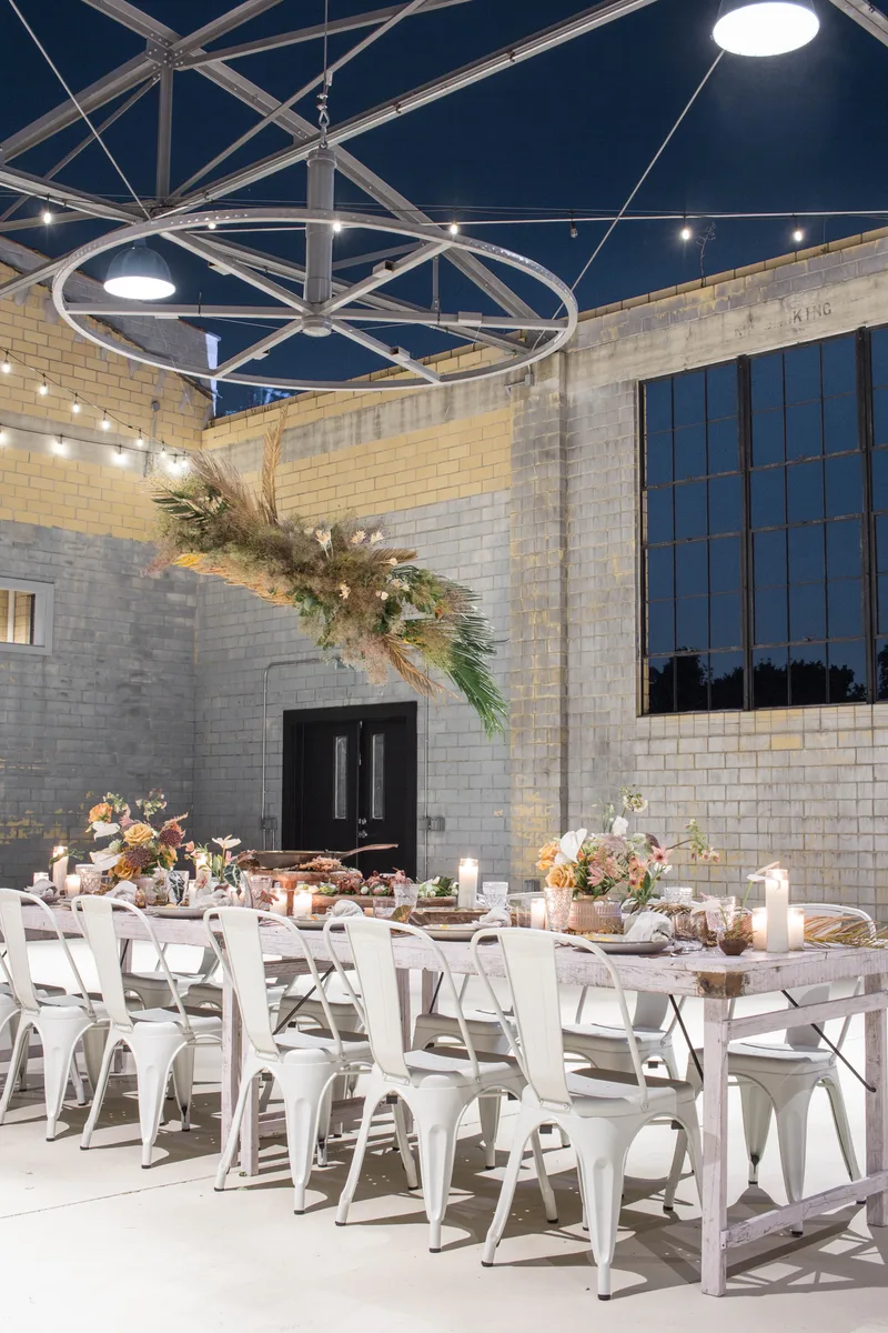 Long candlelit banquet table with white metal chairs under a suspended pampas installation at night