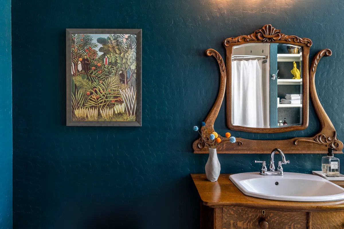 Deep teal bathroom with a carved antique oak vanity, porcelain sink and framed jungle painting