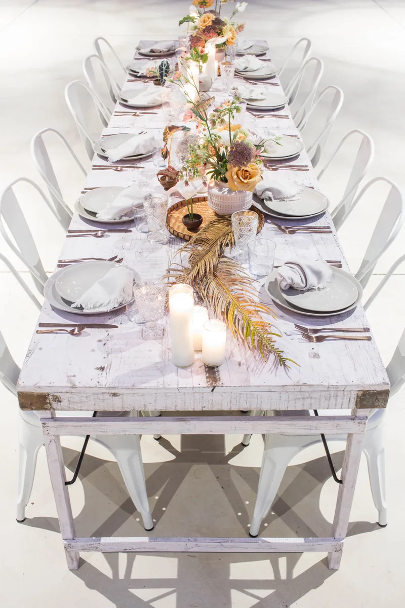 Long whitewashed farm table set with grey plates, candles, dried palm fronds and peach roses