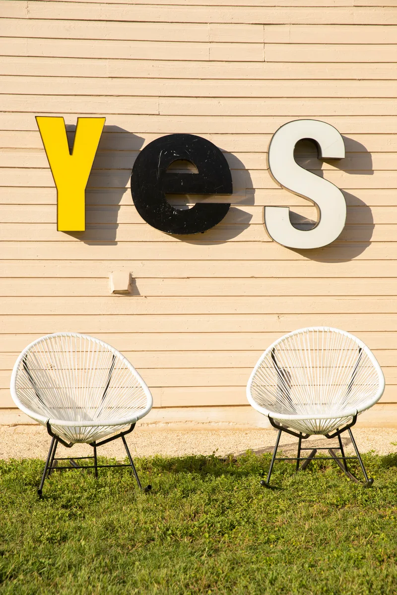 Salvaged marquee letters spelling Yes on a clapboard wall above two white string chairs