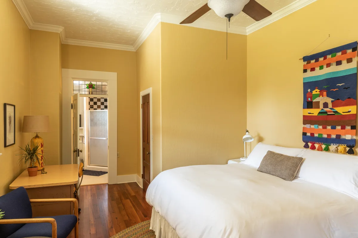 Principal's Office bedroom in golden yellow, white bed, woven wall hanging, bathroom beyond