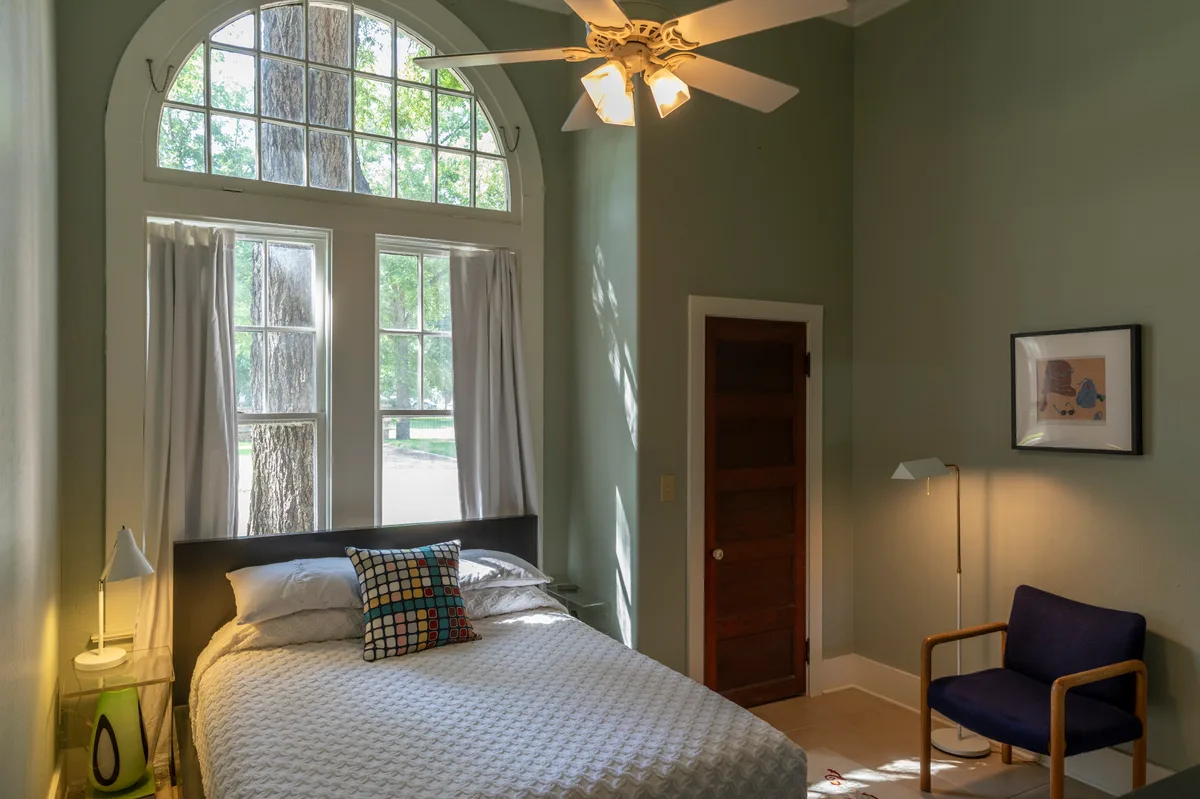 Sage green bedroom beneath a tall arched window with dark wood door and navy armchair