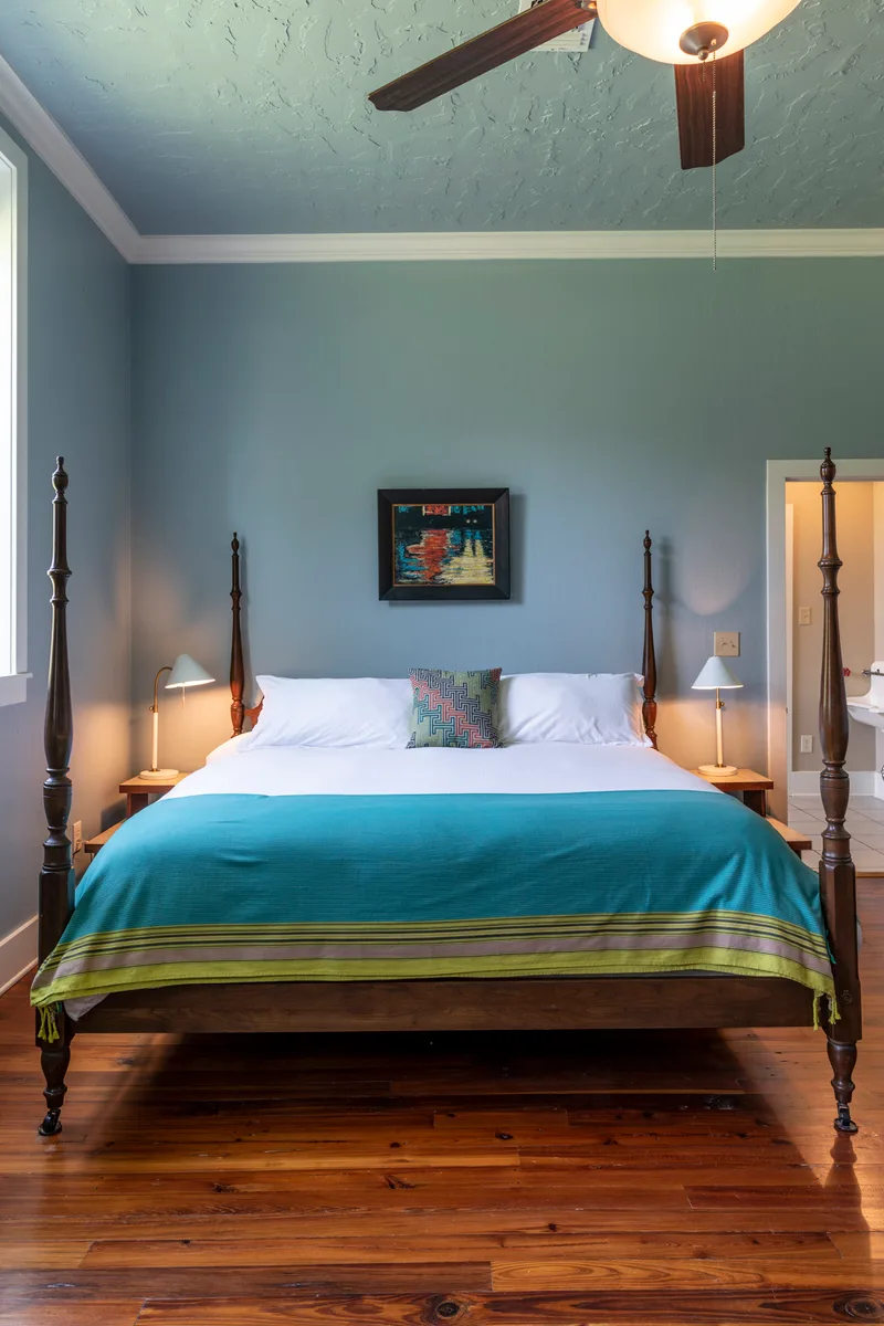 Four-poster bed with teal blanket in a blue-grey bedroom with ceiling fan and wood floors