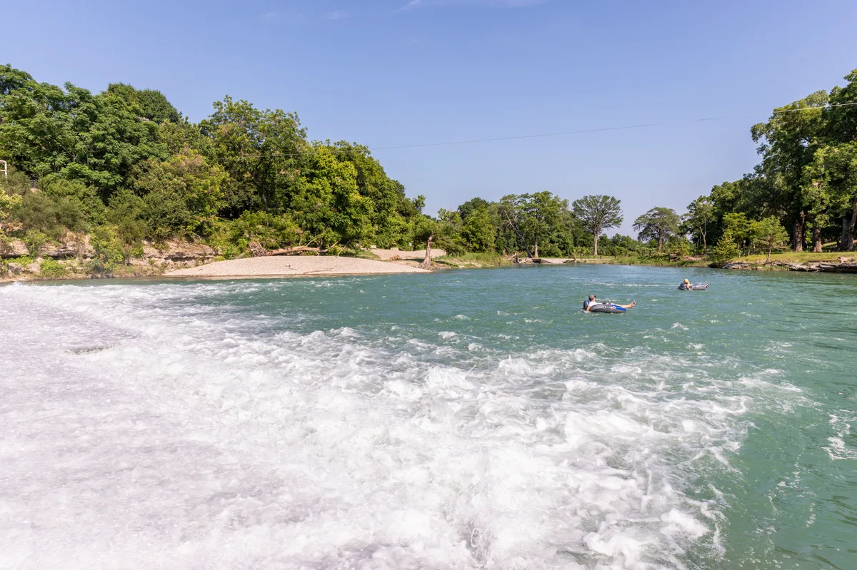 Tubers floating on the green San Marcos River past a gravel bank and whitewater rapids