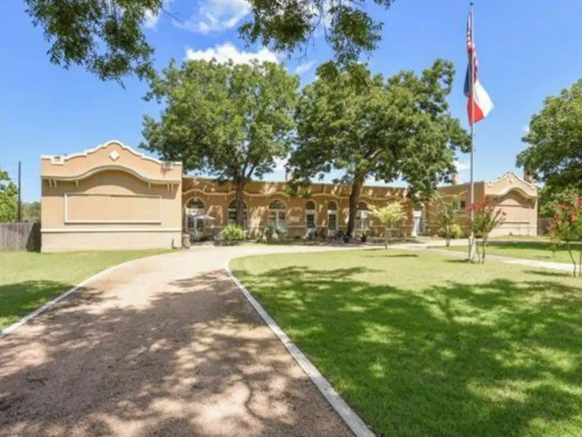 Front of the Schoolhouse, tan stucco building with curved parapets, shade trees and Texas flag