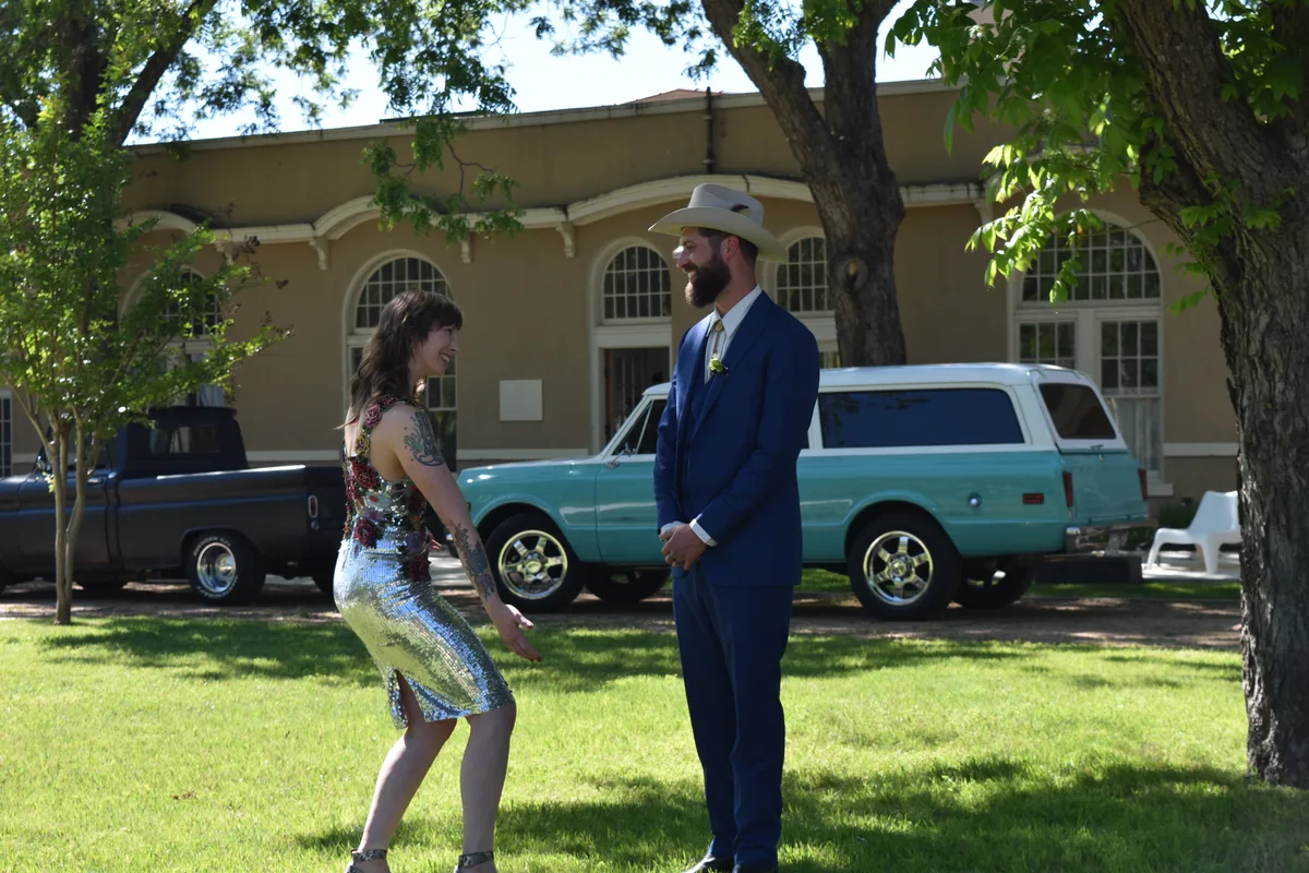 Couple in sequined dress and navy suit laughing on the lawn beside a vintage turquoise truck