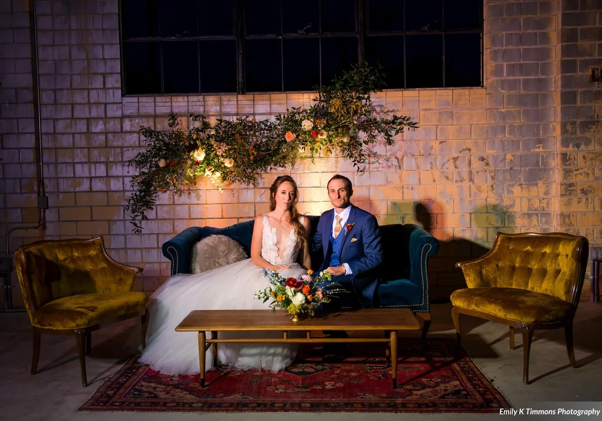 Bride and groom on a teal velvet sofa beneath a lit floral installation, mustard armchairs flanking