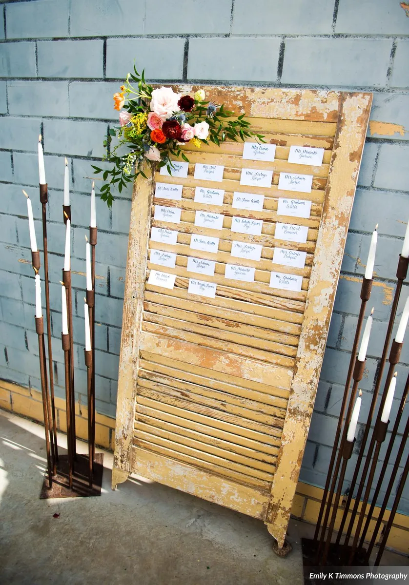 Chipped yellow louvered shutter used as an escort-card display, flanked by tall taper candle stands