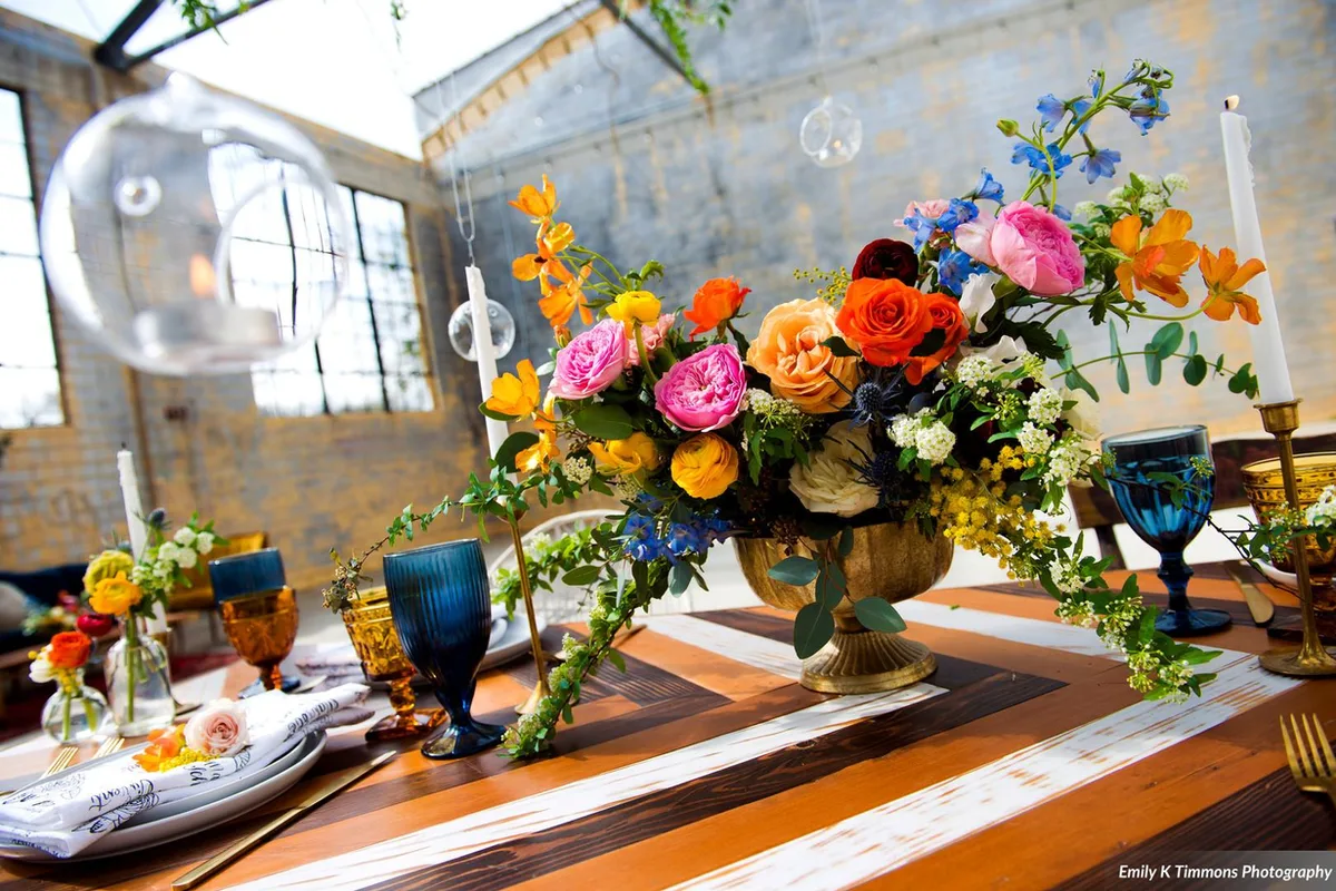 Gold urn centerpiece of pink, orange and blue blooms with amber and cobalt goblets, striped table
