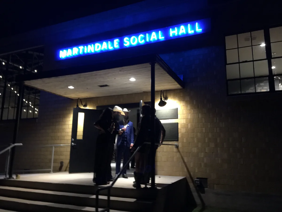 Blue neon Martindale Social Hall sign glowing above guests gathered at the entrance at night