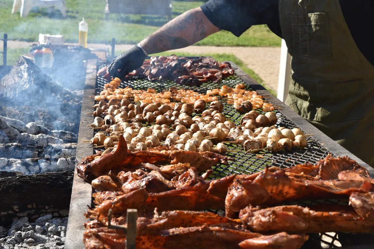 Cook tending a long charcoal grill loaded with skewered mushrooms, shrimp and smoked meats outdoors