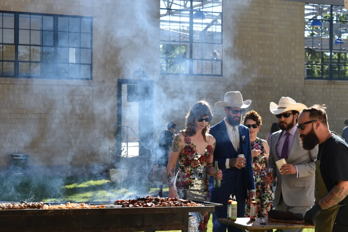 Wedding guests in cowboy hats gather around a smoking barbecue grill outside the Social Hall