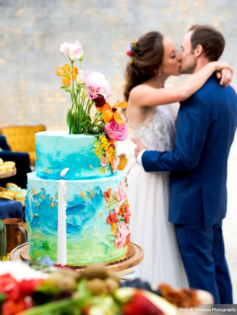 Turquoise ombre buttercream wedding cake topped with bright flowers, a couple kissing blurred behind