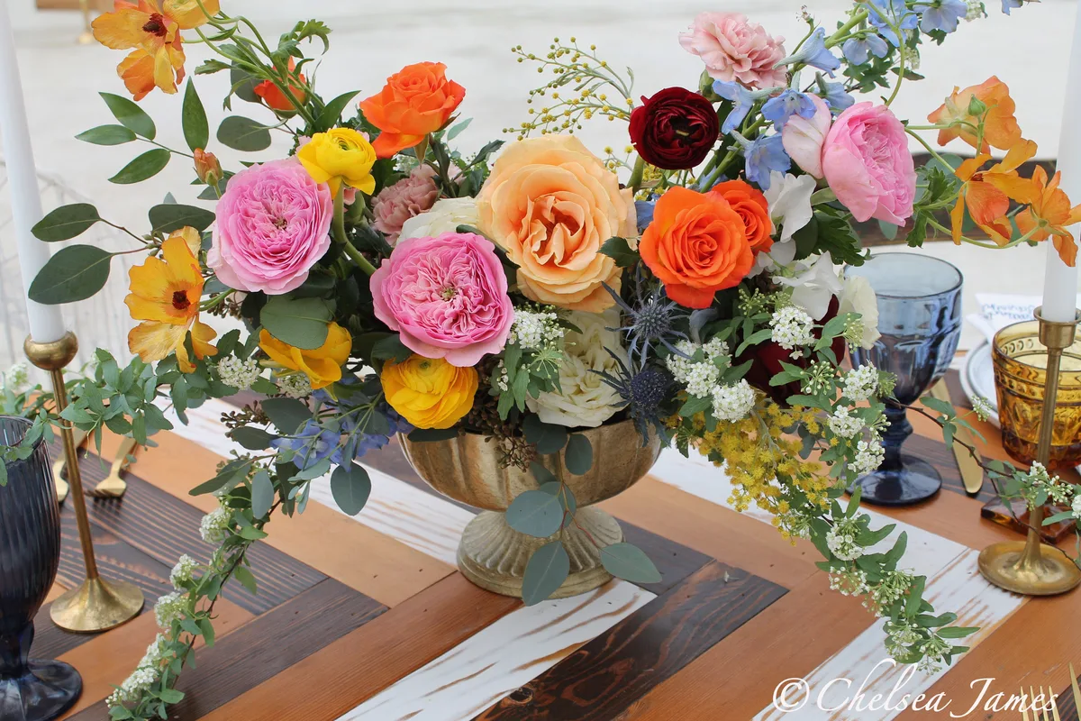 Peach, pink and orange roses in a gold footed bowl beside brass candlesticks and blue goblets