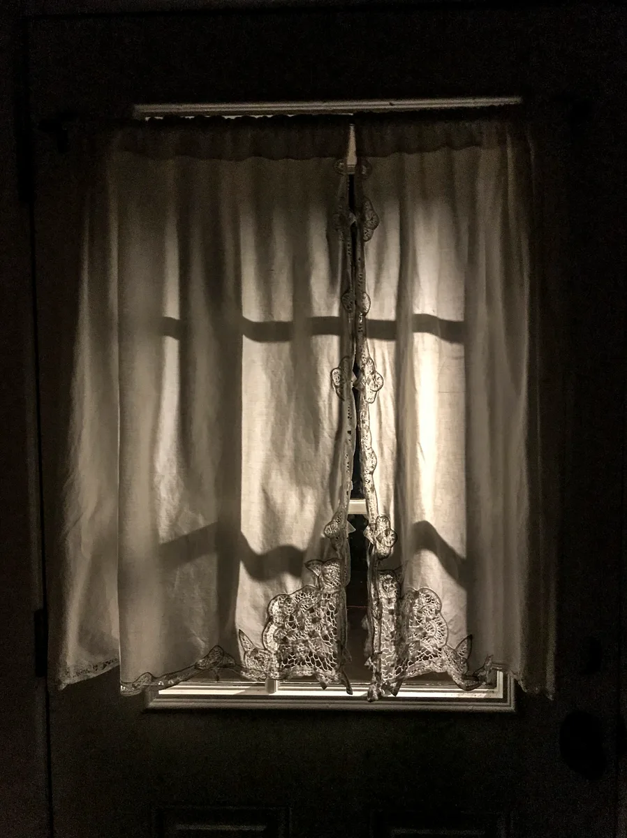 Sheer lace-edged curtains on the principal's office window, a cross-shaped frame shadow falling across them