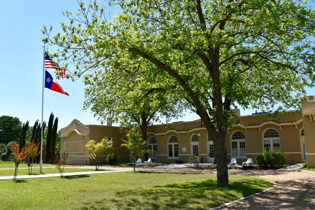 Front of the schoolhouse, tan stucco with tall arched windows, shade tree and flagpole
