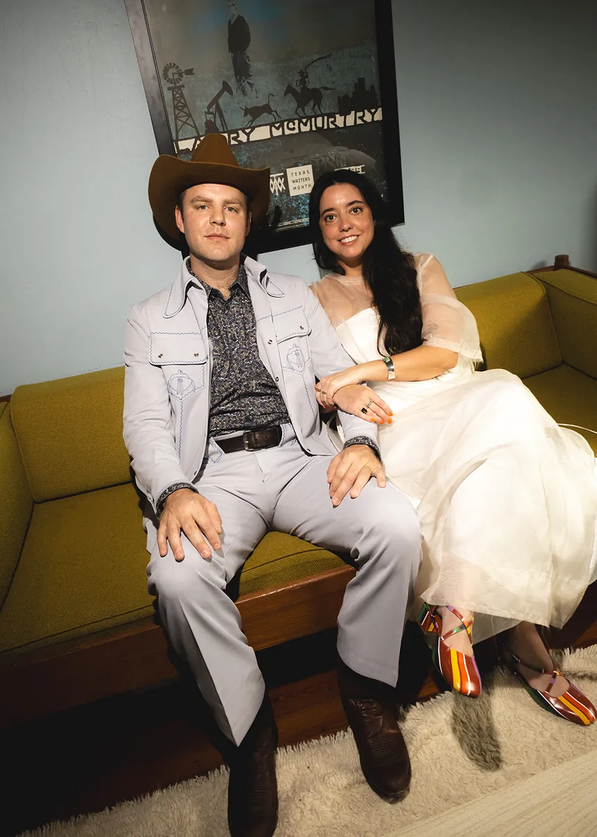 Newlyweds seated together on an olive sofa, the groom in a pale grey western suit and cowboy hat, the bride in white
