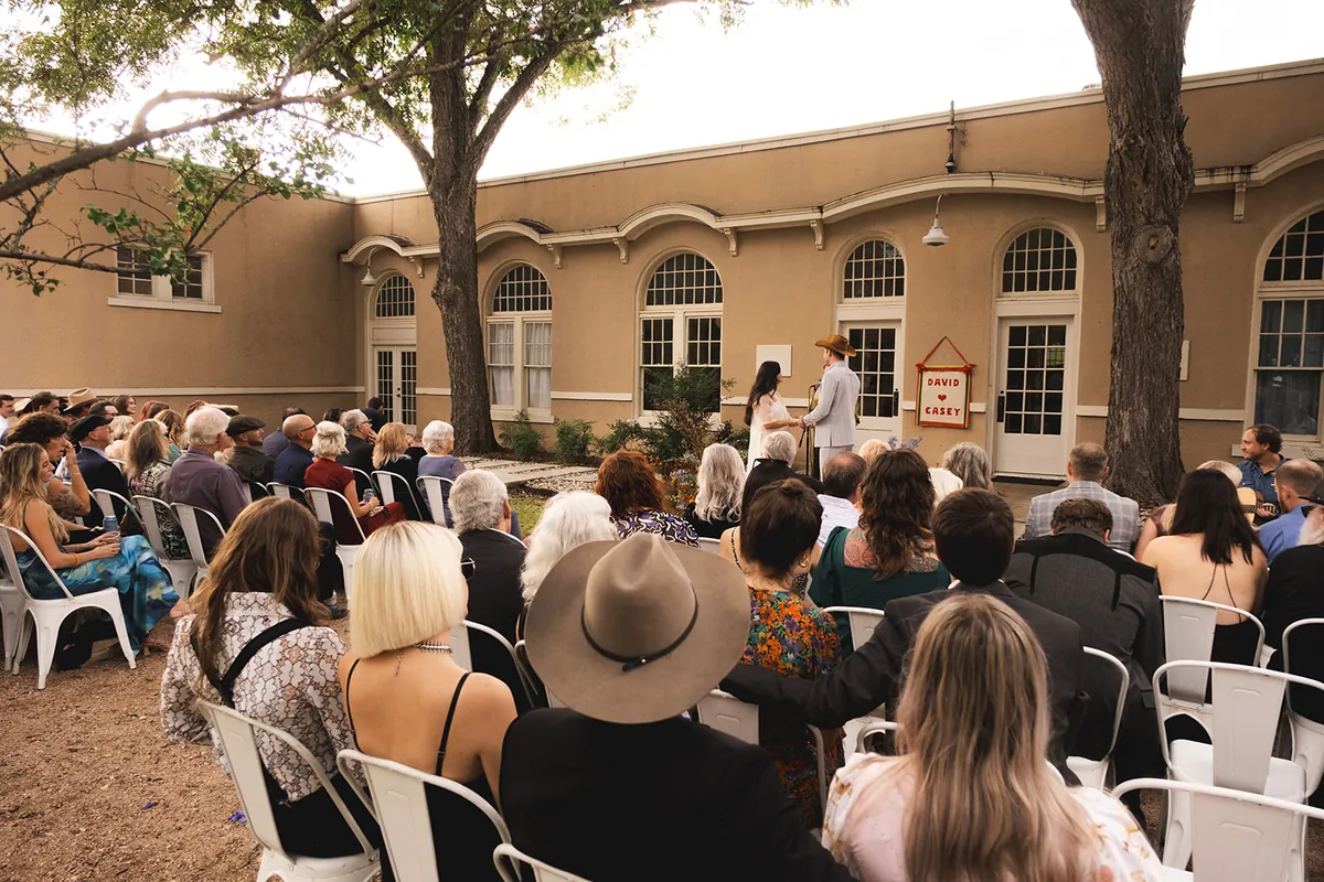 Guests in white chairs watch an outdoor ceremony beneath the trees in front of the Schoolhouse and its arched windows
