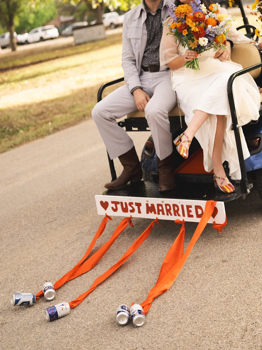 Newlyweds seated on the back of a golf cart with a Just Married sign and cans trailing on orange ribbons