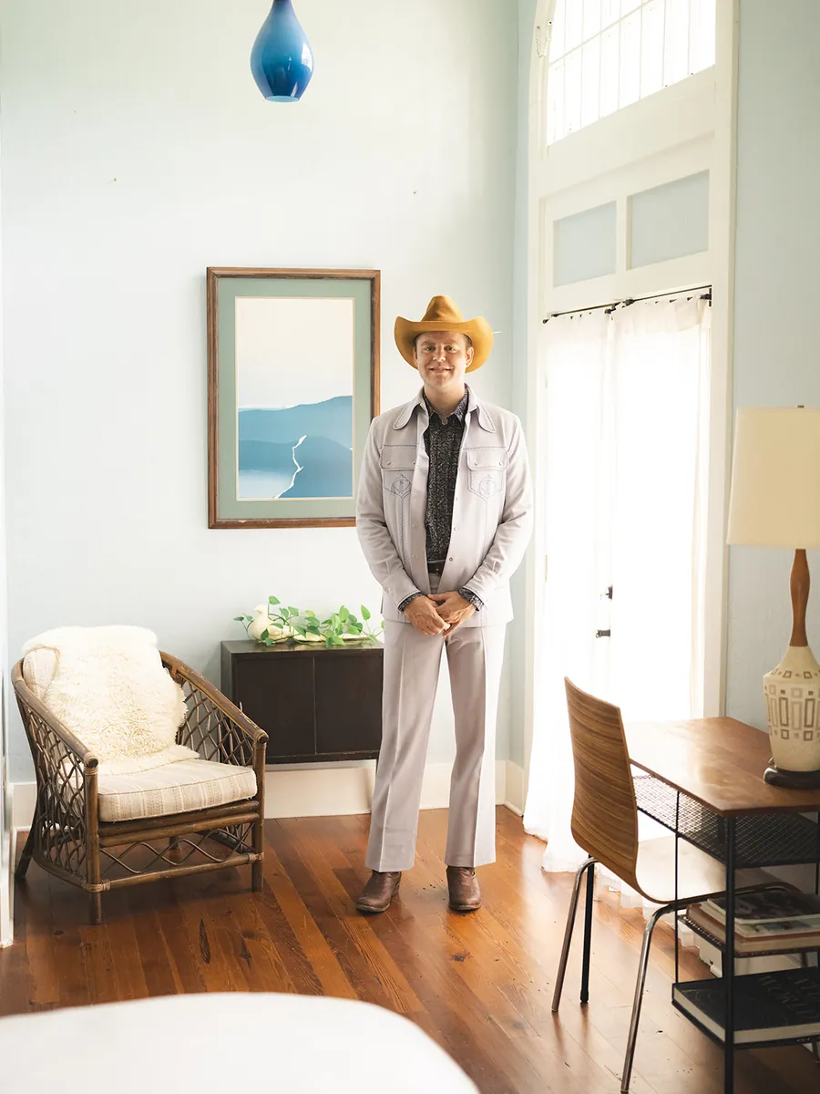 Groom in a pale grey western suit and tan cowboy hat in a sunlit guest room beside a rattan armchair