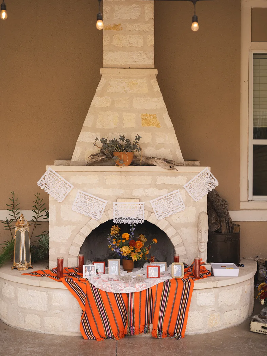 Limestone outdoor fireplace dressed with white papel picado, marigolds, candles and framed photographs on an orange striped blanket