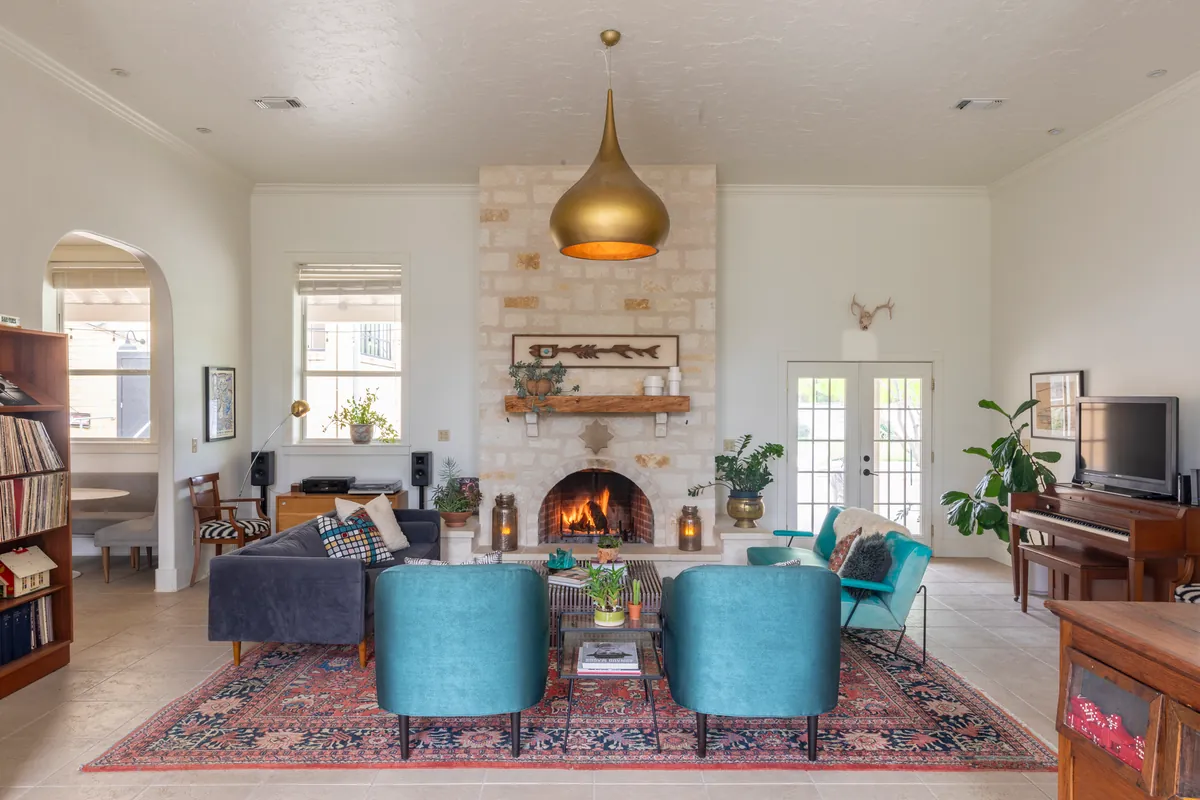 Schoolhouse living room with a lit limestone fireplace, brass teardrop pendant, teal armchairs and a red Persian rug