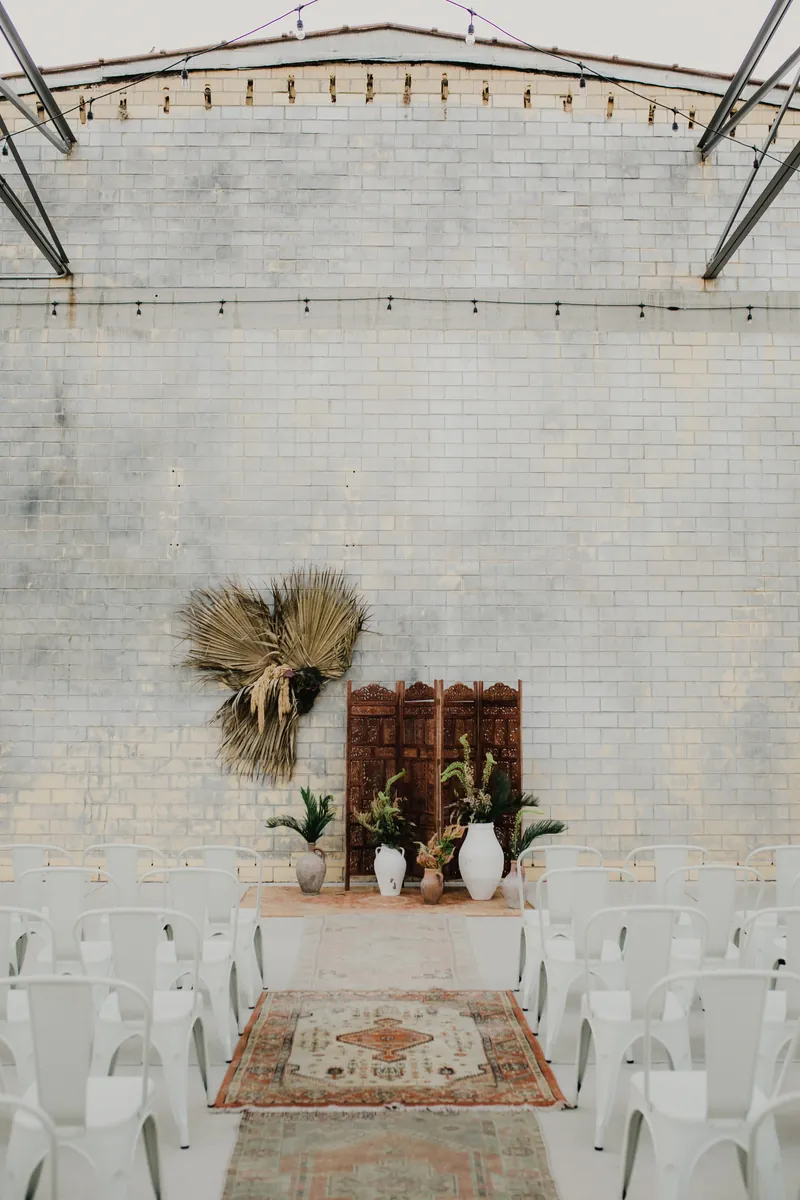 The Social Hall set for a ceremony, white chairs lining a vintage rug aisle to dried palms and a carved wooden screen