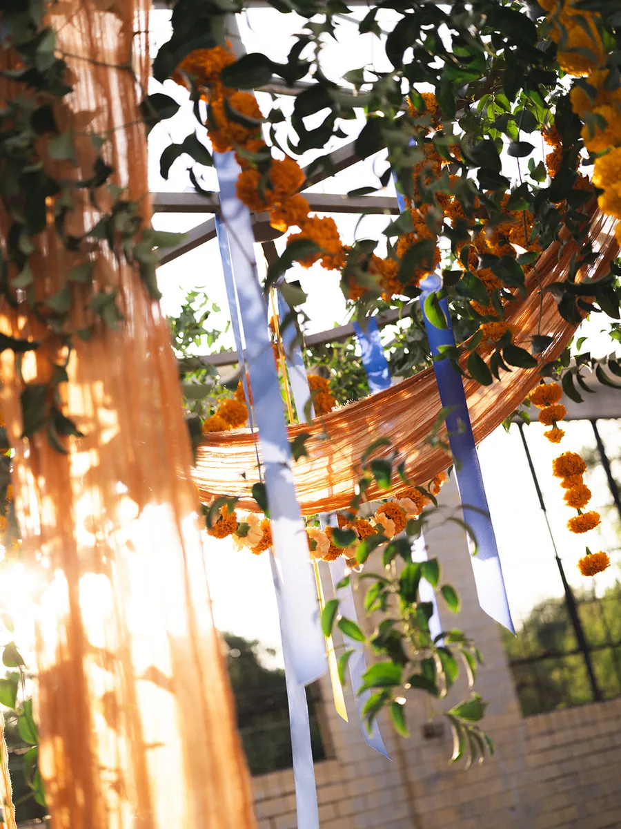 Marigold garlands, trailing greenery, blue ribbons and sheer orange fabric hanging from an overhead frame, backlit by the sun